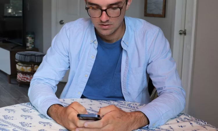 Một người đàn ông mặc áo sơ mi xanh đang ngồi ở bàn và gõ trên điện thoại thông minh